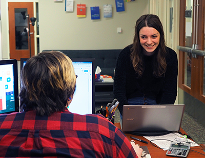 a young woman approaches a desk. Another young woman is facing her and has computer screens open